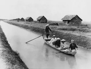 War Victims Prints: Vietnamese men in a sampan on a canal near straw dwellings that house refugees from North Vietnam