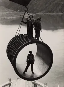 Washington State Prints: Four workers riding on a large casing section of pipe suspended from cable as it is moved into position during